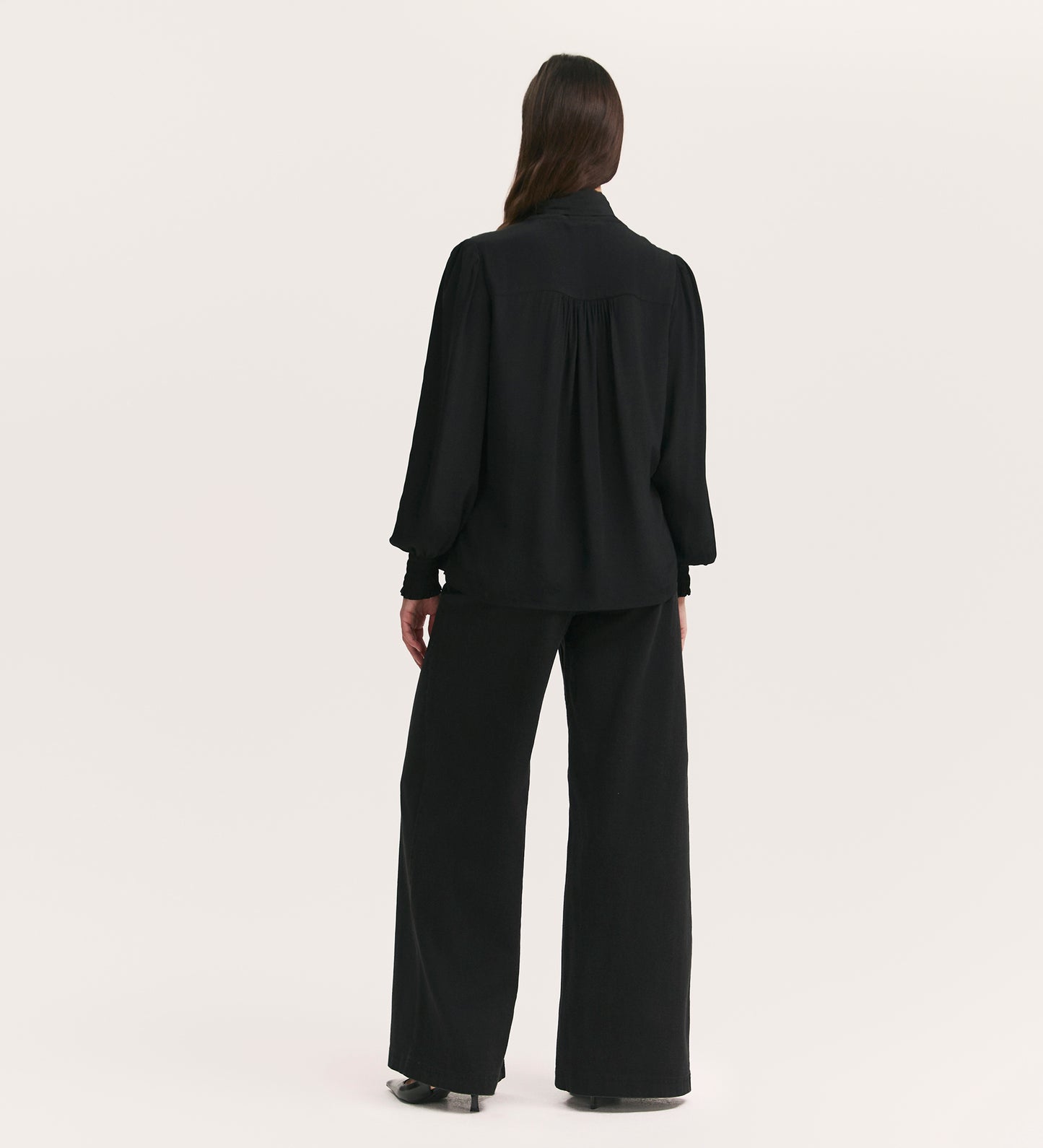 A person with long, dark hair stands facing away, wearing the Mavis Black Chiffon Blouse with long sleeves and a loose fit, paired with wide-leg black pants against a plain light background.