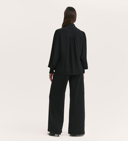 A person with long, dark hair stands facing away, wearing the Mavis Black Chiffon Blouse with long sleeves and a loose fit, paired with wide-leg black pants against a plain light background.