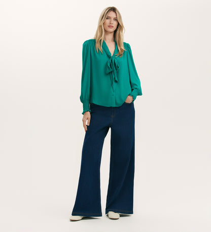 A woman stands against a plain background wearing the Mavis Turquoise Chiffon Blouse, paired with wide-leg dark blue jeans and white shoes.