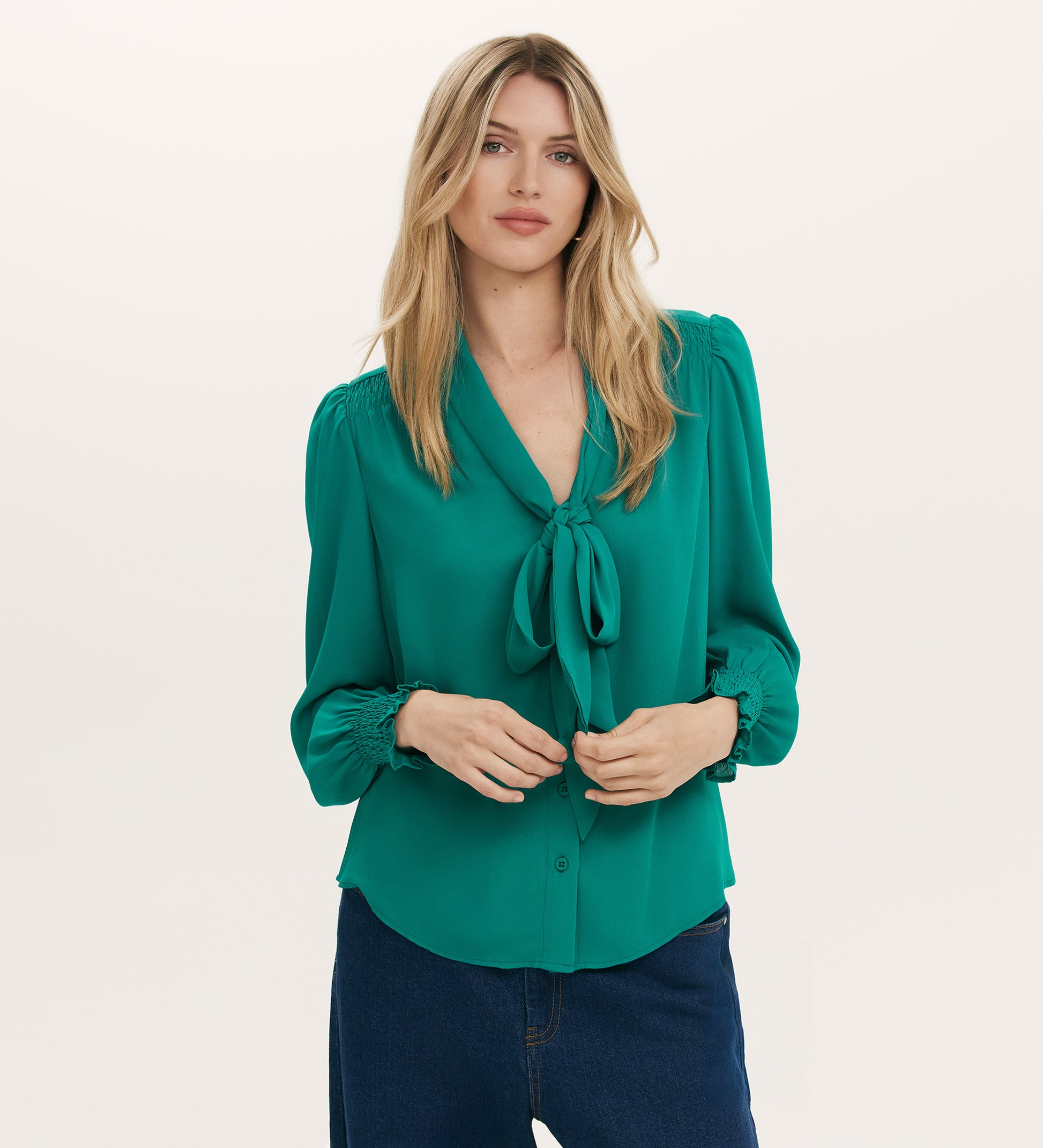 A woman with long blonde hair wears the Mavis Turquoise Chiffon Blouse with smocked cuffs, paired with dark blue jeans, standing against a plain light background.