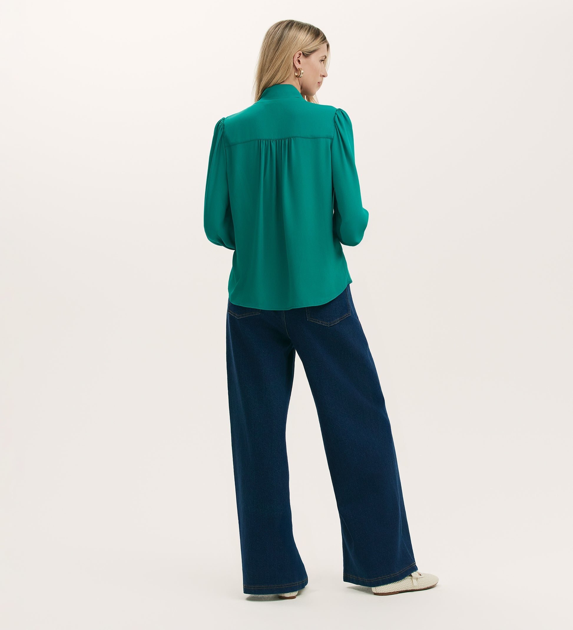 A woman with long blonde hair faces away, wearing the Mavis Turquoise Chiffon Blouse paired with wide-leg dark blue jeans and white shoes, set against a plain light background.