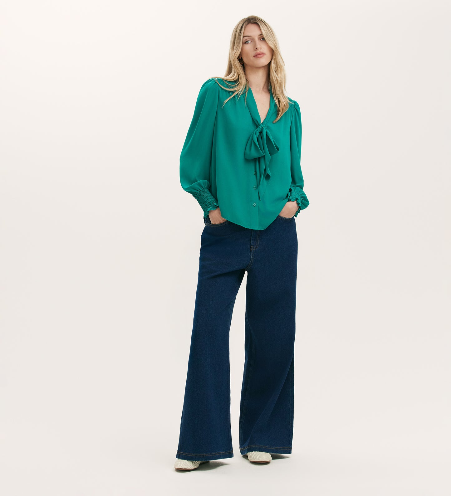 A woman with long blonde hair wears the Mavis Turquoise Chiffon Blouse with long sleeves, paired with wide-leg dark blue jeans and white shoes, standing against a plain background to highlight the blouse's elegant versatility.