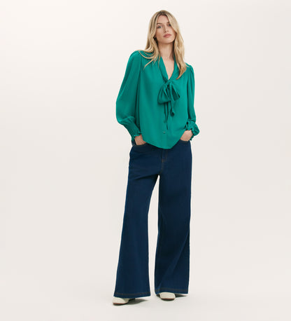 A woman with long blonde hair wears the Mavis Turquoise Chiffon Blouse with long sleeves, paired with wide-leg dark blue jeans and white shoes, standing against a plain background to highlight the blouse's elegant versatility.