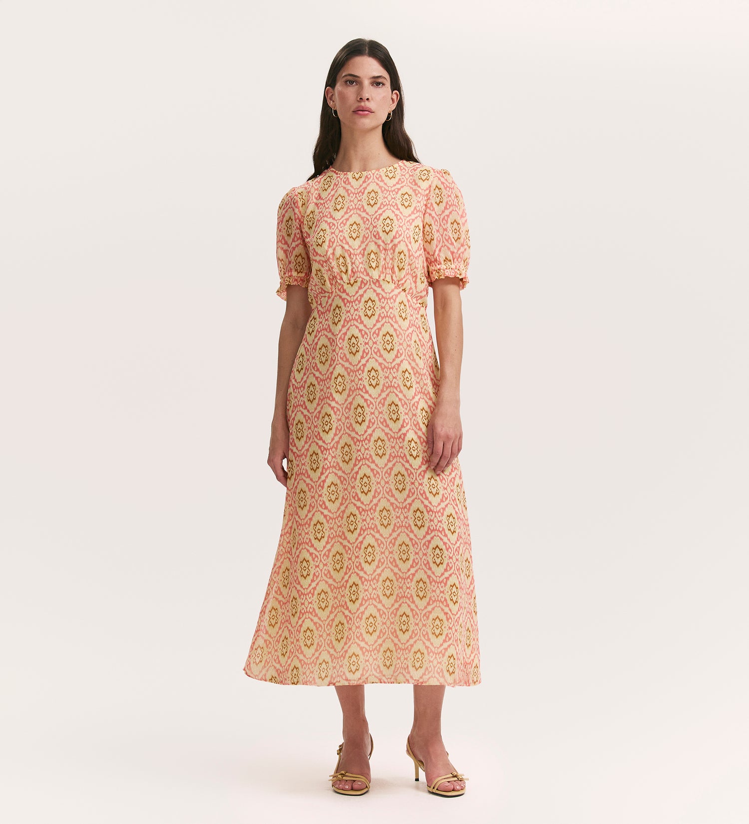 A woman wears the Mela Coral Ikat Chiffon Midi Tea Dress, featuring short sleeves, a fitted waist, and a flowing skirt, styled with gold heeled sandals against a plain light background.