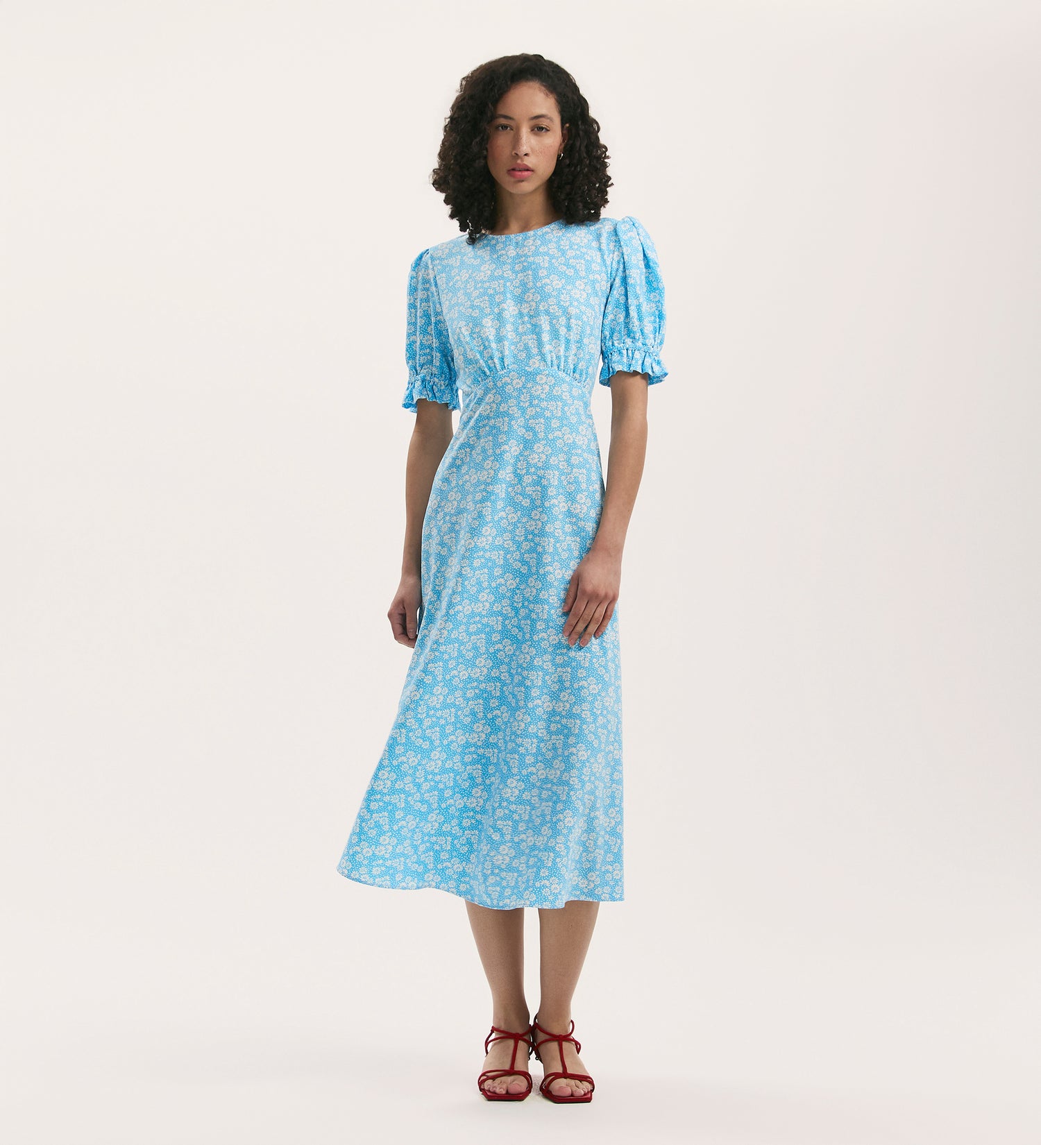 A woman with curly hair wears the Mela Blue Daisy Crepe Midi Tea Dress, featuring puffed short sleeves and an empire line waist, paired with red strappy sandals against a plain light background.