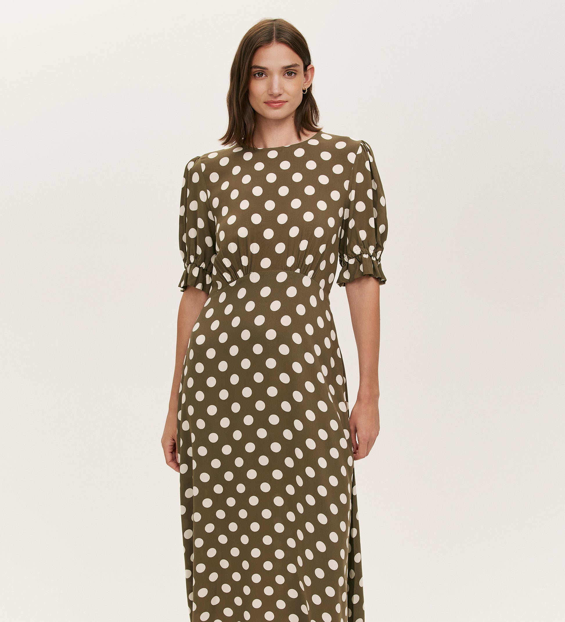 A woman stands against a plain light background wearing the Mela Khaki Polka Dot Midi Dress, featuring white polka dots, short puffed sleeves, and a flattering empire line waist.