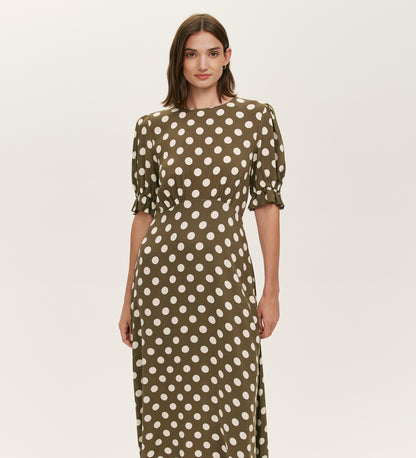 A woman stands against a plain light background wearing the Mela Khaki Polka Dot Midi Dress, featuring white polka dots, short puffed sleeves, and a flattering empire line waist.
