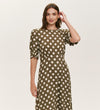 A woman in the Mela Khaki Polka Dot Midi Dress, featuring white spots, short sleeves, and a fitted waist, stands against a plain light background gently touching her hair.