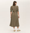 A woman faces away, wearing the Mela Khaki Polka Dot Midi Dress with an empire waist and short puff sleeves, paired with white socks and yellow sandals, set against a plain light background.