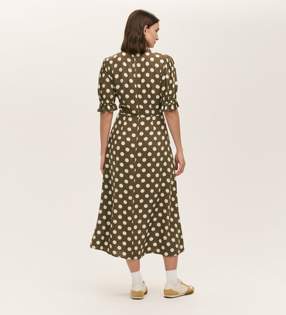 A woman faces away, wearing the Mela Khaki Polka Dot Midi Dress with an empire waist and short puff sleeves, paired with white socks and yellow sandals, set against a plain light background.