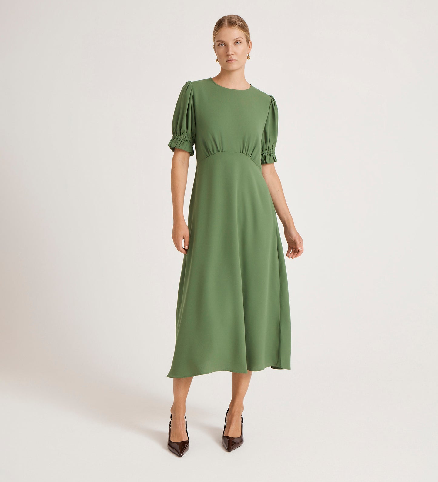 A woman in the Mela Green Chiffon Midi Tea Dress stands against a plain white background. Her hair is pulled back and she wears black pointed-toe heels.