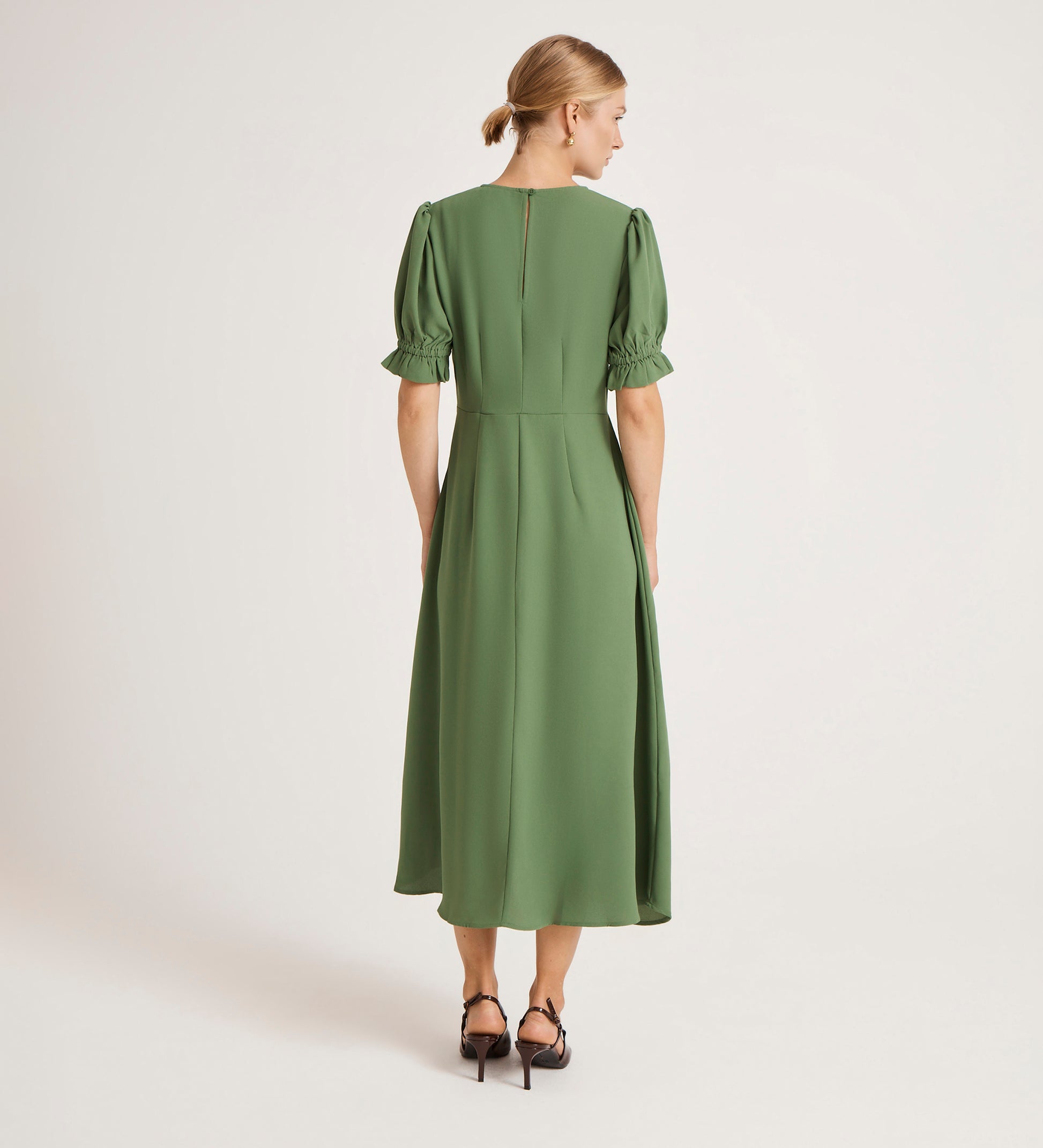 A woman with blonde hair in a low bun is seen from behind wearing the Mela Green Chiffon Midi Tea Dress with puffed short sleeves and black heeled sandals, standing against a plain light background.