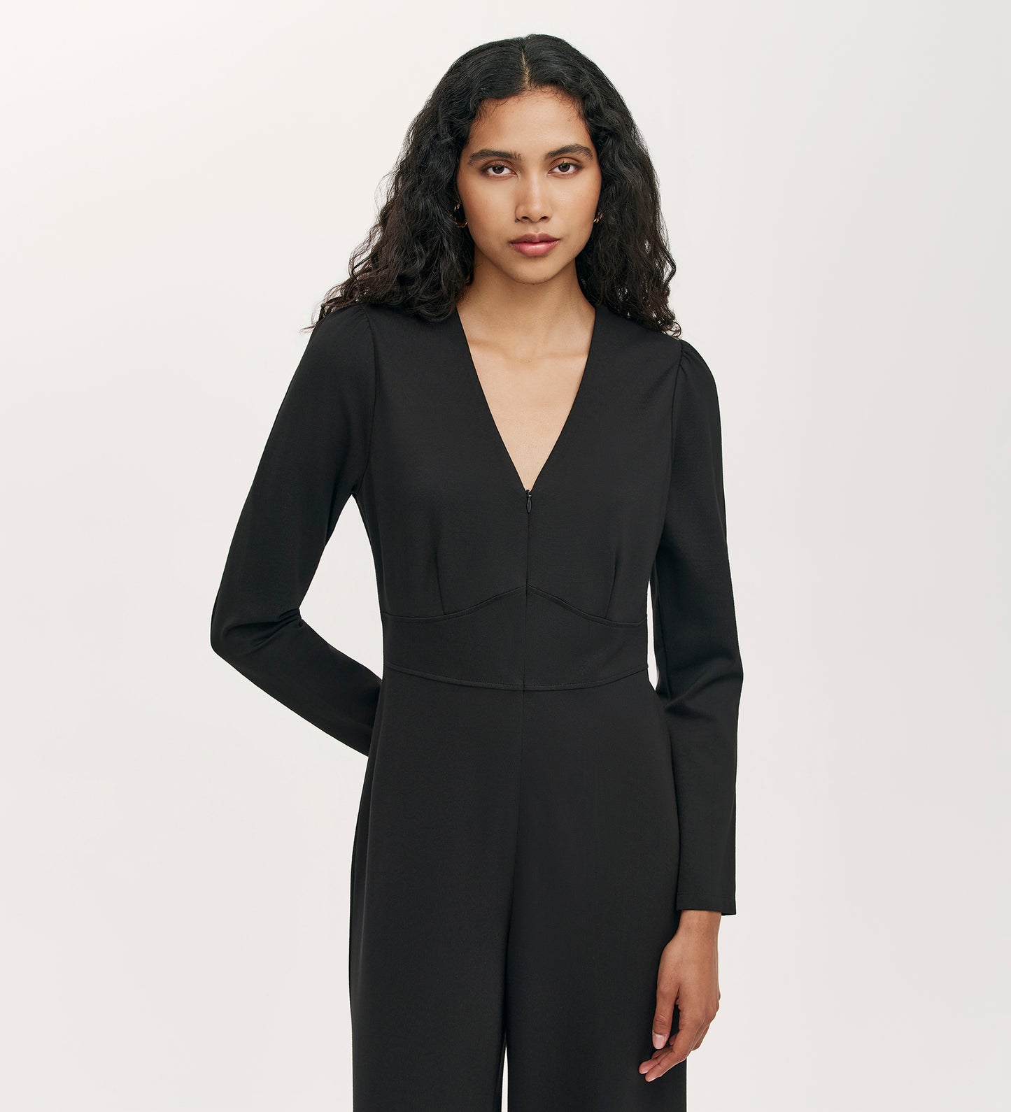 A woman with long, wavy dark hair wears the Melly Black Ponte Jersey Jumpsuit—black, long-sleeved with wide-leg trousers—standing against a plain light background with a neutral expression.