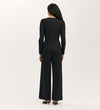 A woman faces away, wearing the Melly Black Ponte Jersey Jumpsuit with wide-leg trousers and black heeled sandals against a plain, light-colored background.
