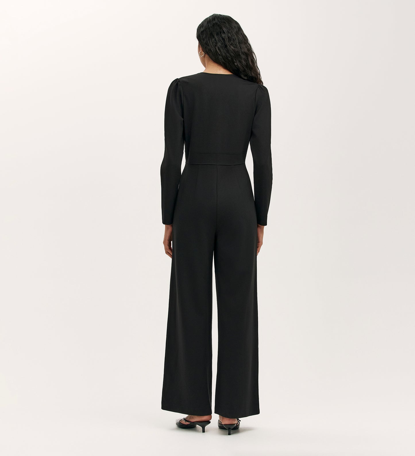 A woman faces away, wearing the Melly Black Ponte Jersey Jumpsuit with wide-leg trousers and black heeled sandals against a plain, light-colored background.