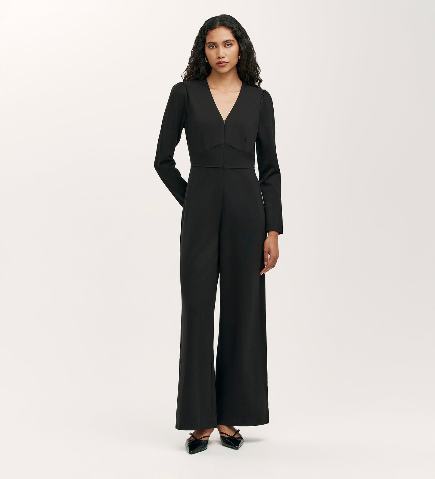 A woman with long curly hair wears the Melly Black Ponte Jersey Jumpsuit, featuring long sleeves, a V-neck, and wide-leg trousers, paired with black pointed-toe flats against a plain light background.