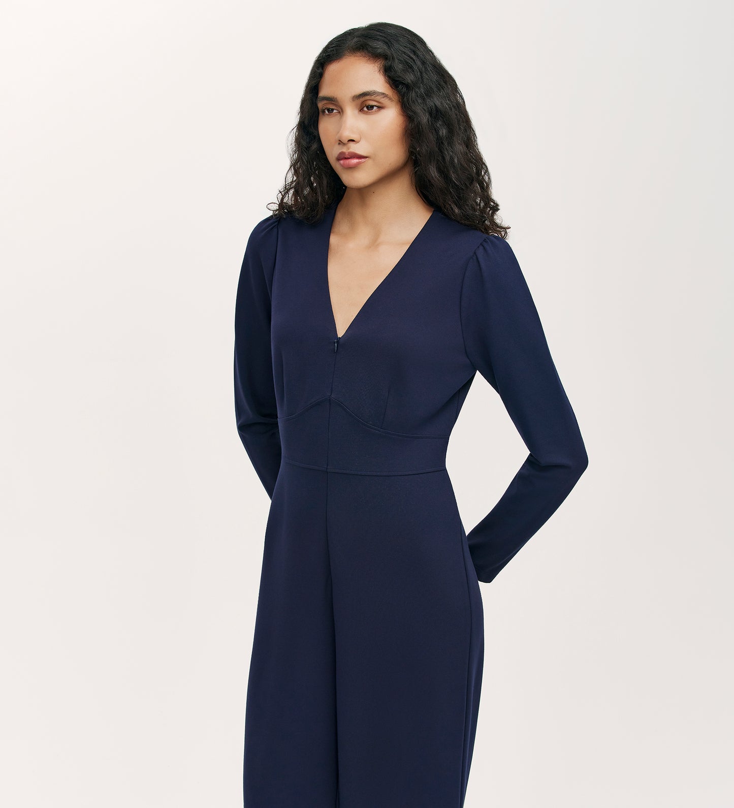 A woman with long curly hair stands against a plain light background, looking to the side with her hands behind her back, wearing the Melly Navy Ponte Jersey Jumpsuit featuring wide-leg trousers.