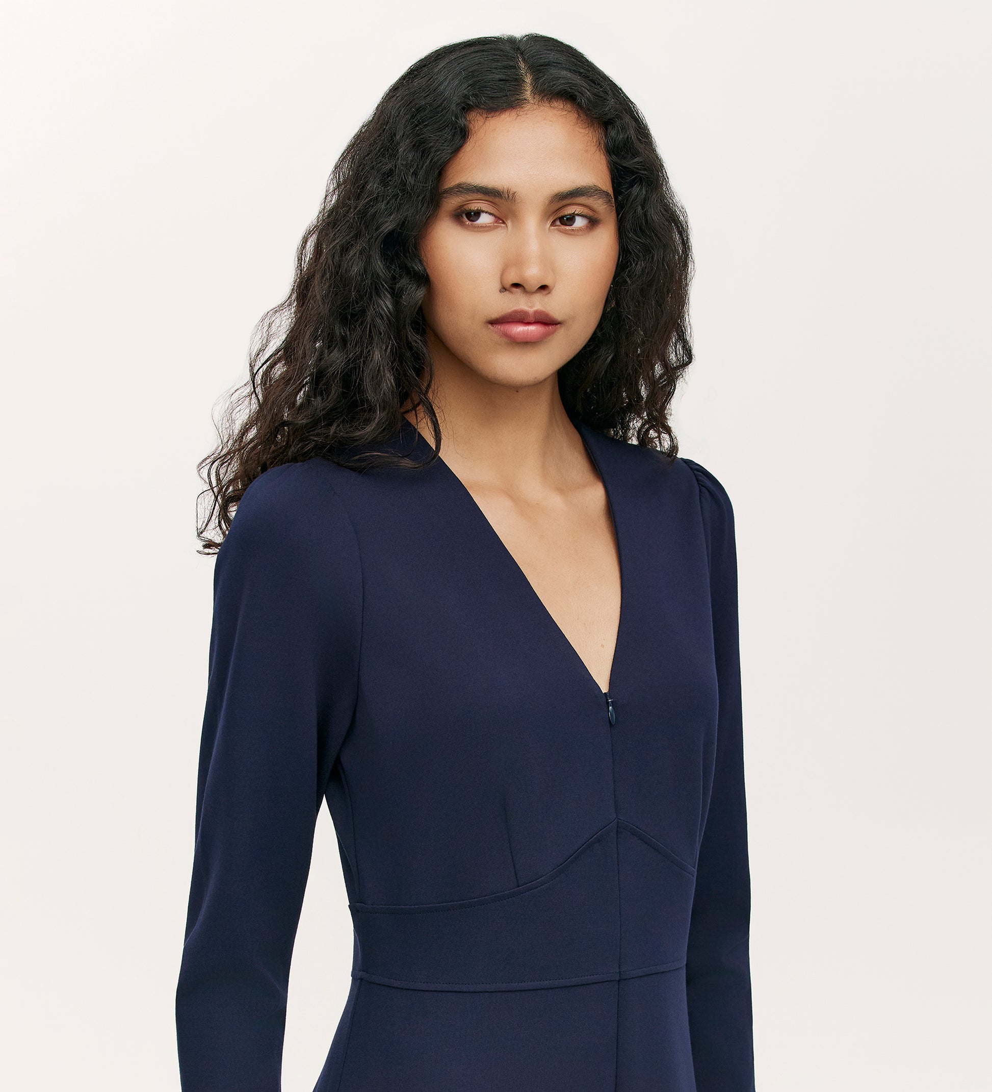 A woman with long, wavy dark hair wearing the Melly Navy Ponte Jersey Jumpsuit looks slightly to the side against a plain light background.