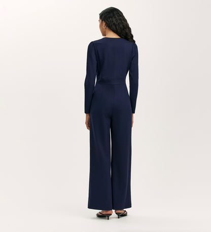 A woman with long wavy hair, shown from the back, wears the Melly Navy Ponte Jersey Jumpsuit with wide-leg trousers and black heels, standing against a plain light background.