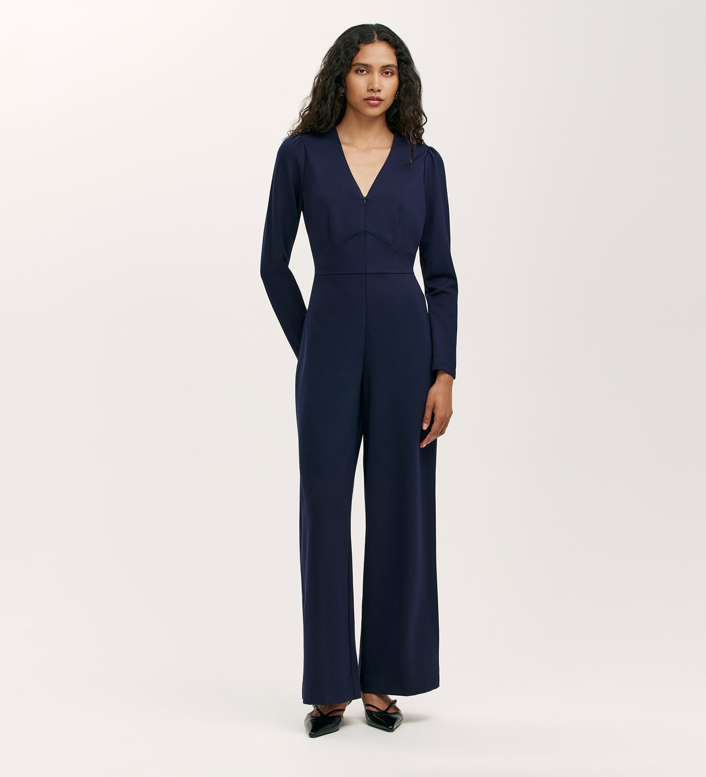 A woman with long, wavy hair wears the Melly Navy Ponte Jersey Jumpsuit with a V-neckline, long sleeves, and wide-leg trousers, standing against a plain light background and pairing it with black pointed shoes.
