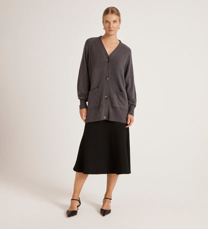 A woman stands against a plain background wearing the Mila Charcoal Knitted Cardigan with large buttons, a V-neckline, and front pockets, paired with a black midi skirt and black pointed-toe heels.