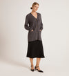 A woman stands against a plain background wearing the Mila Charcoal Knitted Cardigan styled over a black midi skirt and ankle-strap shoes. Her hands are in the cardigan pockets, and her hair is tied back.