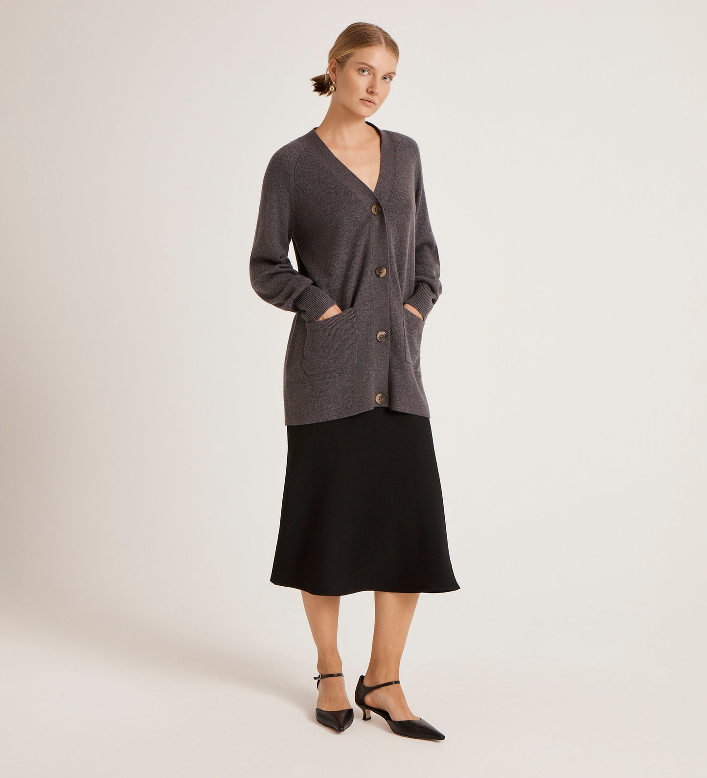 A woman stands against a plain background wearing the Mila Charcoal Knitted Cardigan styled over a black midi skirt and ankle-strap shoes. Her hands are in the cardigan pockets, and her hair is tied back.