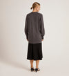 A woman with her hair in a low bun faces away, wearing the Mila Charcoal Knitted Cardigan over a black midi skirt and black heeled sandals against a plain, light background.