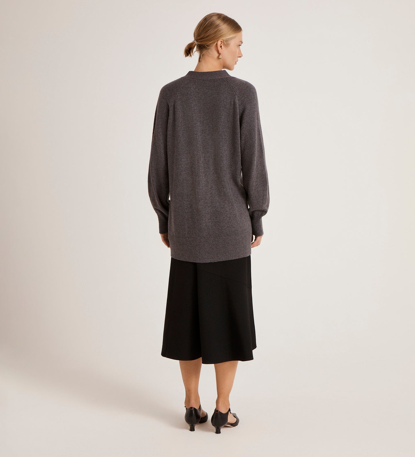 A woman with her hair in a low bun faces away, wearing the Mila Charcoal Knitted Cardigan over a black midi skirt and black heeled sandals against a plain, light background.