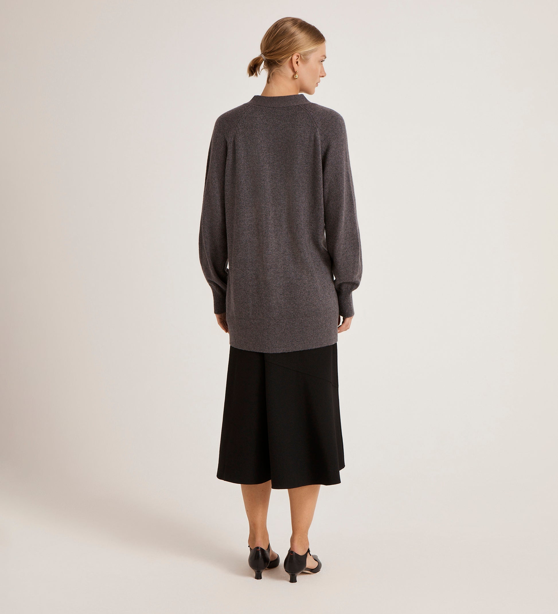 A woman with her hair in a low bun faces away, wearing the Mila Charcoal Knitted Cardigan over a black midi skirt and black heeled sandals against a plain, light background.