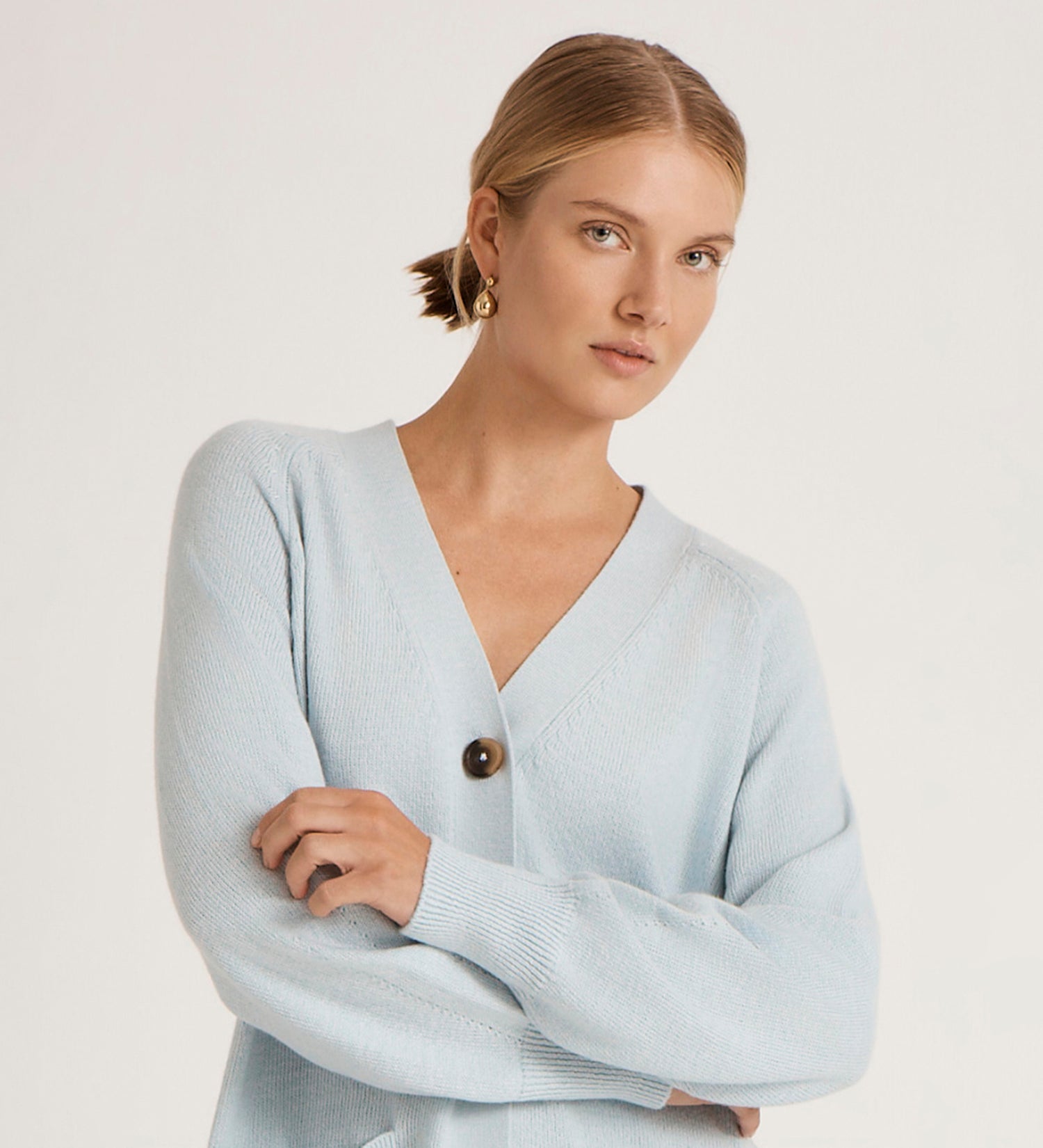 A woman with light hair pulled back wears the Mila Light Blue Knitted Cardigan—an oversized, chunky knit with a V-neckline and large dark button—paired with gold earrings against a plain, light background.