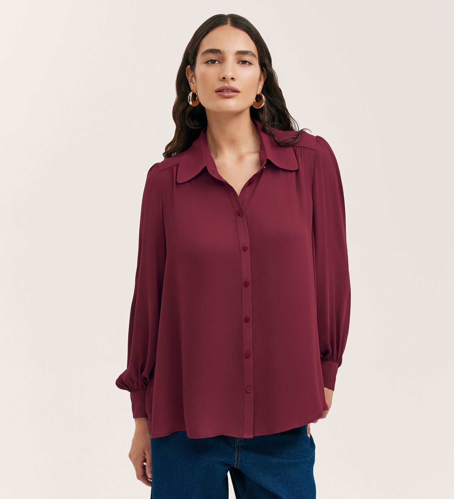 A woman stands against a plain light background wearing the Melina Burgundy Crepe Blouse, a relaxed fit, long-sleeve button-up with a collar, paired with blue jeans.