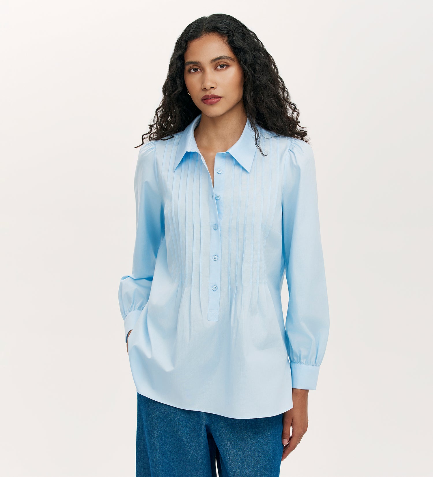 A woman with long, wavy dark hair wears the Mimi Blue Cotton Rich Shirt, a blue button-up with pintuck detail and long sleeves. She stands with her hands in her pockets against a plain light background.