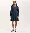 A woman stands against a plain light background wearing the Nancy Indigo Denim Dress, a relaxed-fit mini with long sleeves and a tiered skirt. Her loose, wavy hair complements her look, paired with white socks and blue sneakers. She gazes ahead neutrally.