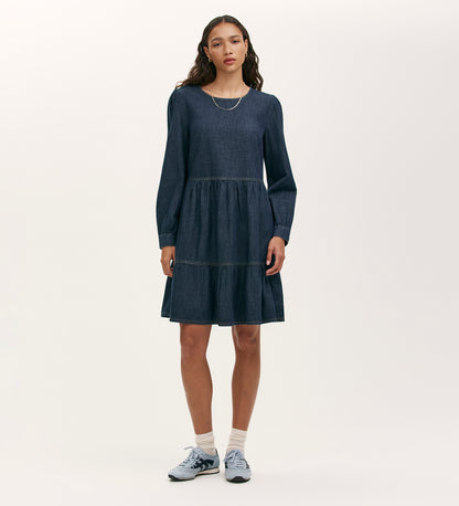 A woman stands against a plain light background wearing the Nancy Indigo Denim Dress, a relaxed-fit mini with long sleeves and a tiered skirt. Her loose, wavy hair complements her look, paired with white socks and blue sneakers. She gazes ahead neutrally.
