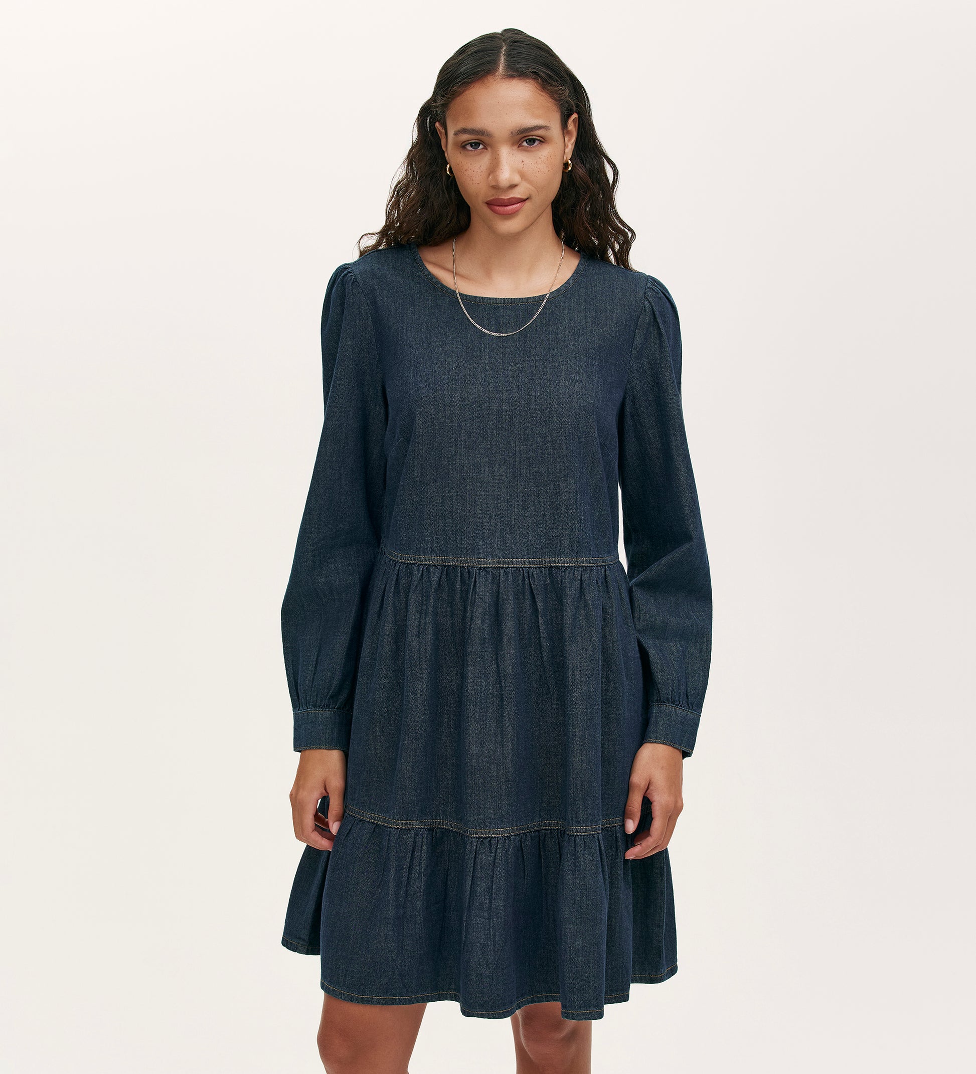 A woman with long, wavy hair wears the Nancy Indigo Denim Dress, featuring a relaxed fit, long sleeves, and a tiered mini skirt. She pairs it with a simple necklace and stands against a plain light background.