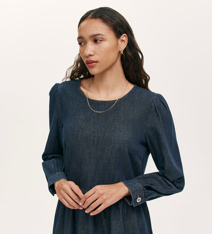 A woman with long, wavy brown hair wears the Nancy Indigo Denim Dress with gold hoop earrings and a thin gold necklace, standing against a plain light background and looking slightly to the side with her hands gently clasped.