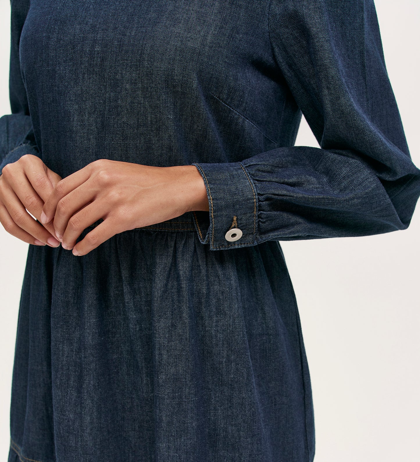 Model in the Nancy Indigo Denim Dress, a long-sleeved dark denim mini with buttoned cuffs, shown from shoulders to mid-thigh, hands clasped at the waist against a plain background.