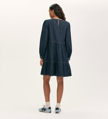 A woman stands facing away in the Nancy Indigo Denim Dress, a relaxed-fit, long-sleeved, knee-length dark denim piece with tiered skirt. She pairs it with white socks and blue sneakers against a light background.