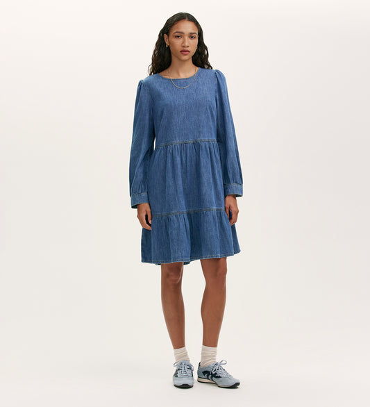 A woman wears the Nancy Light Blue Denim Dress, a relaxed fit, long-sleeve piece with tiered details, styled with white socks and light blue sneakers against a plain light background.