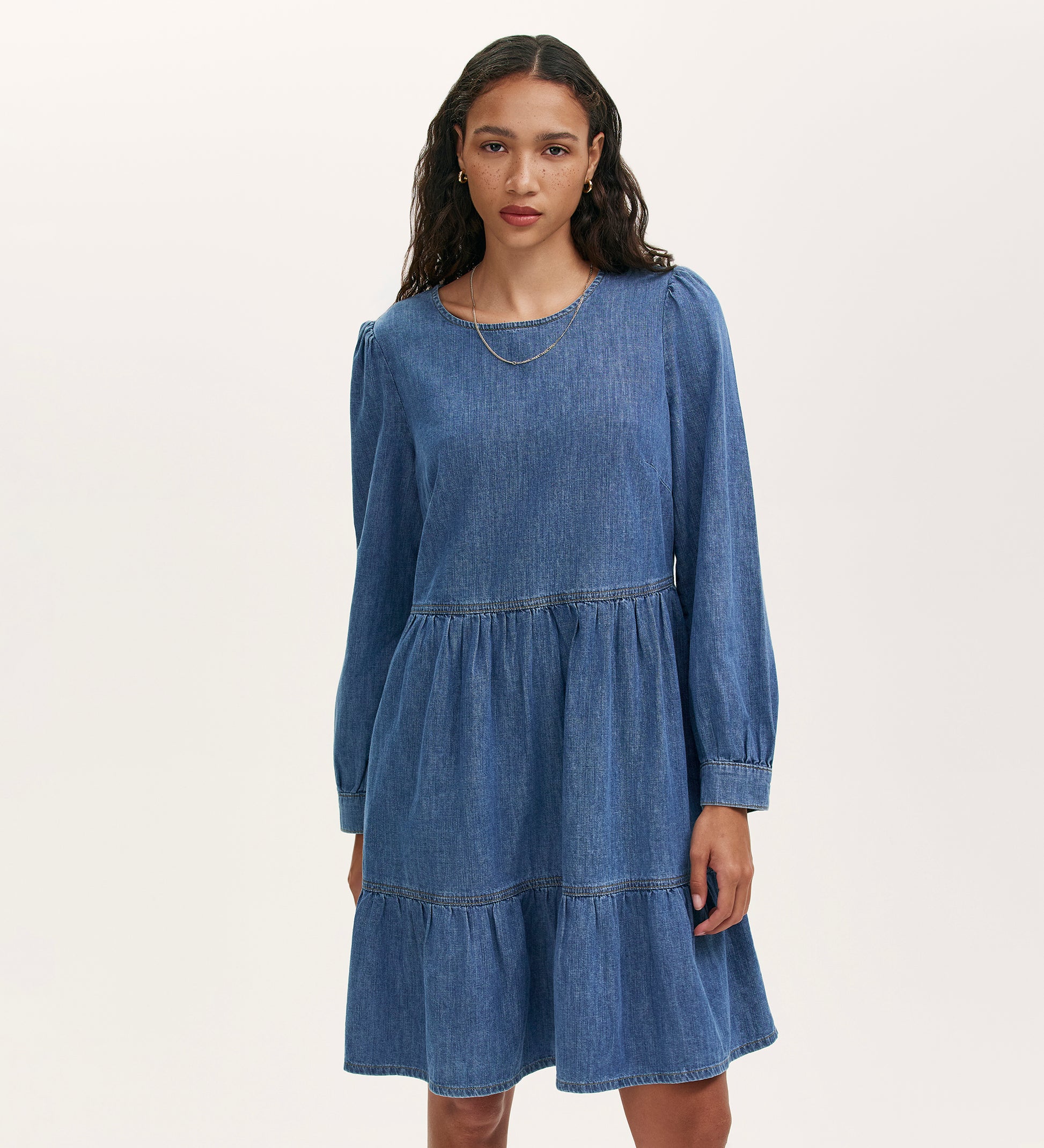 A woman with wavy dark hair wears the Nancy Light Blue Denim Dress, which features long sleeves, a tiered skirt, and subtle puff shoulders. She stands against a plain light background.