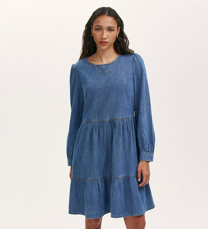 A woman with wavy dark hair wears the Nancy Light Blue Denim Dress, which features long sleeves, a tiered skirt, and subtle puff shoulders. She stands against a plain light background.