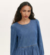 A woman with long, wavy dark hair models the Nancy Light Blue Denim Dress—a light-wash, tiered mini dress—paired with a thin necklace as she stands against a plain light background, looking directly at the camera with a neutral expression.