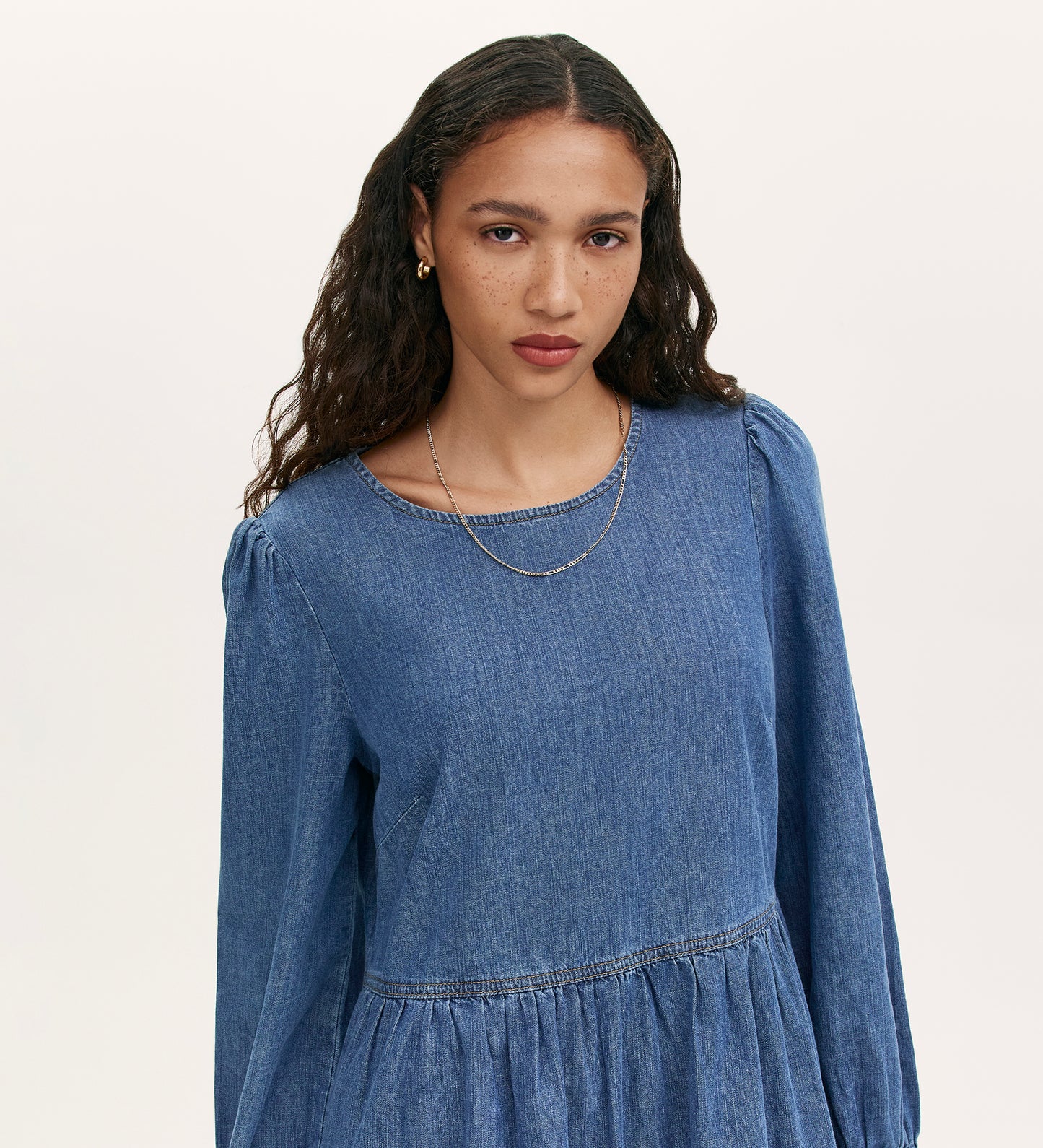 A woman with long, wavy dark hair models the Nancy Light Blue Denim Dress—a light-wash, tiered mini dress—paired with a thin necklace as she stands against a plain light background, looking directly at the camera with a neutral expression.