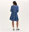 A woman faces away wearing the Nancy Light Blue Denim Dress, a relaxed fit with long sleeves and a tiered skirt, styled with white socks and blue sneakers against a neutral background.