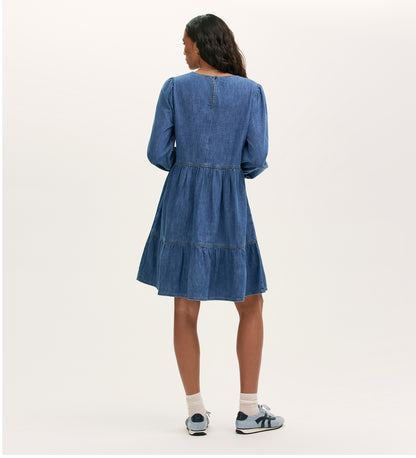 A woman faces away wearing the Nancy Light Blue Denim Dress, a relaxed fit with long sleeves and a tiered skirt, styled with white socks and blue sneakers against a neutral background.