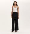 A woman stands facing forward, wearing a white sleeveless tank top, Nellie Black Canvas Wide Leg Trousers with side buttons, and brown heeled ankle boots against a plain light background—a versatile wardrobe staple.