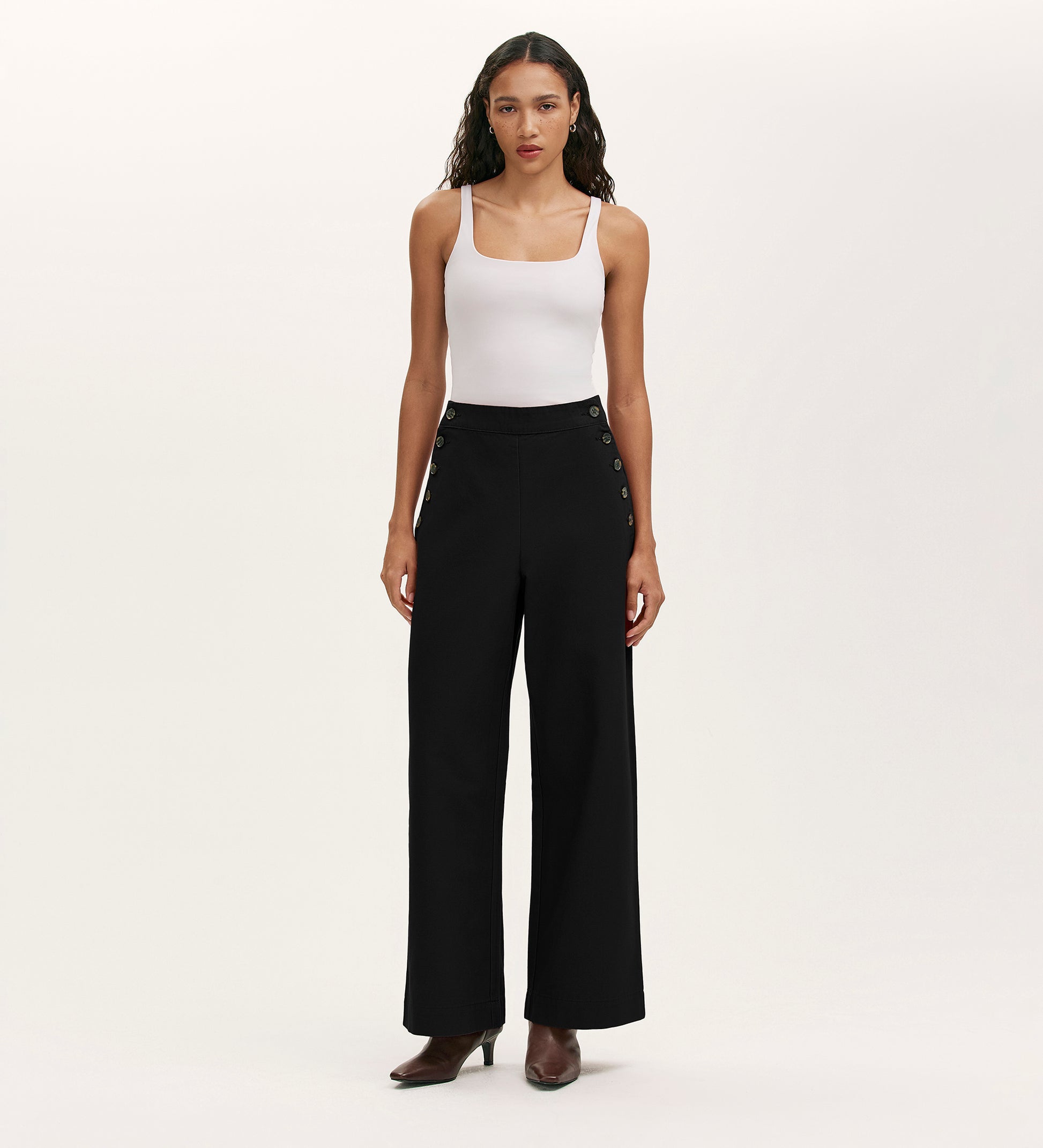 A woman stands facing forward, wearing a white sleeveless tank top, Nellie Black Canvas Wide Leg Trousers with side buttons, and brown heeled ankle boots against a plain light background—a versatile wardrobe staple.