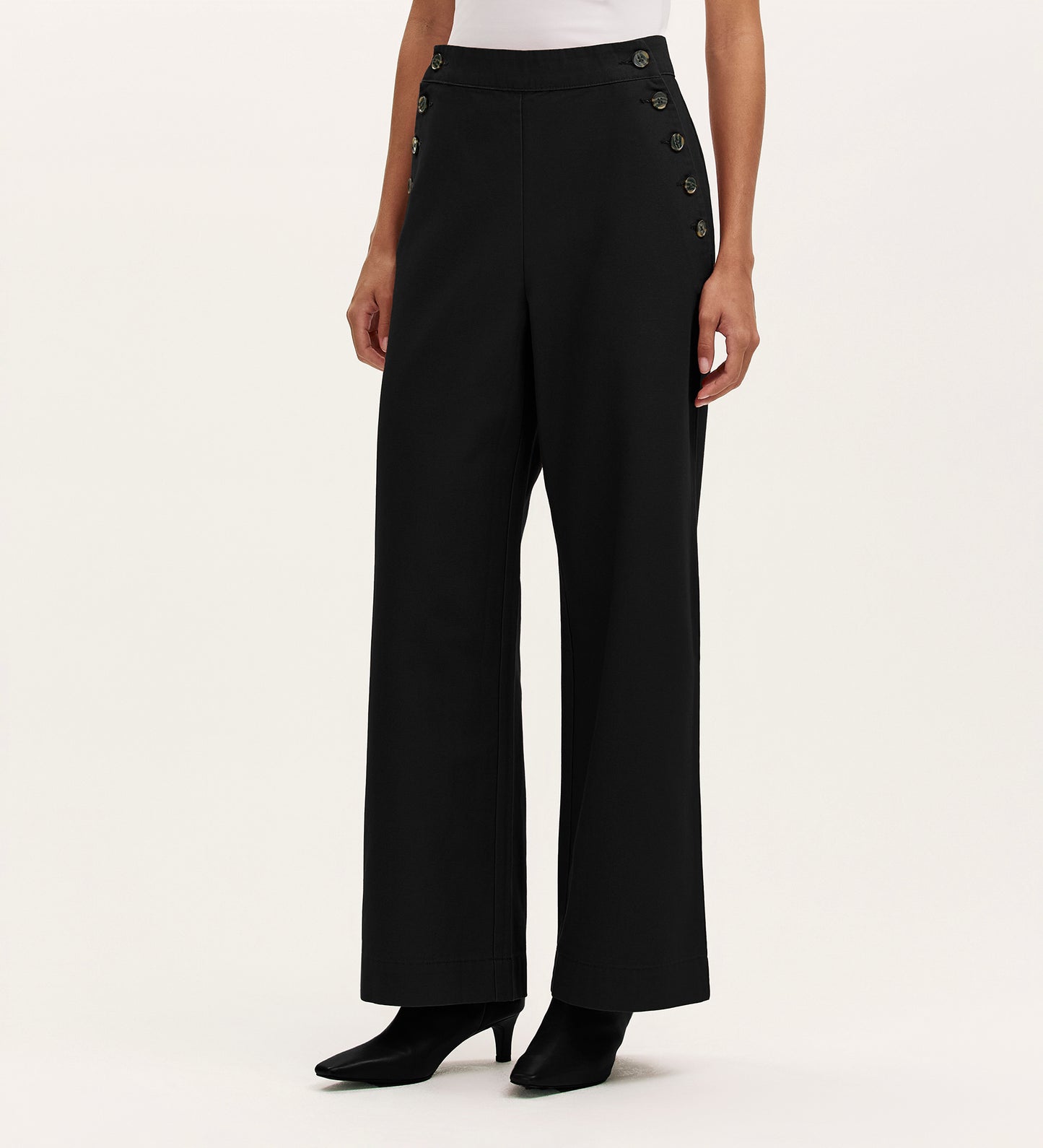 A person wears the Nellie Black Canvas Wide Leg Trousers, featuring high-waisted and side button details, paired with a white top and black heeled boots against a plain light background—showcasing a versatile wardrobe staple.