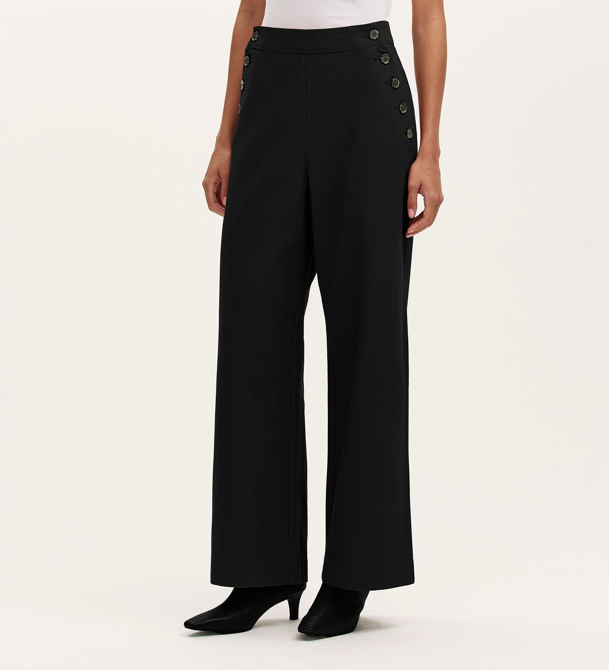 A person wears the Nellie Black Canvas Wide Leg Trousers, featuring high-waisted and side button details, paired with a white top and black heeled boots against a plain light background—showcasing a versatile wardrobe staple.